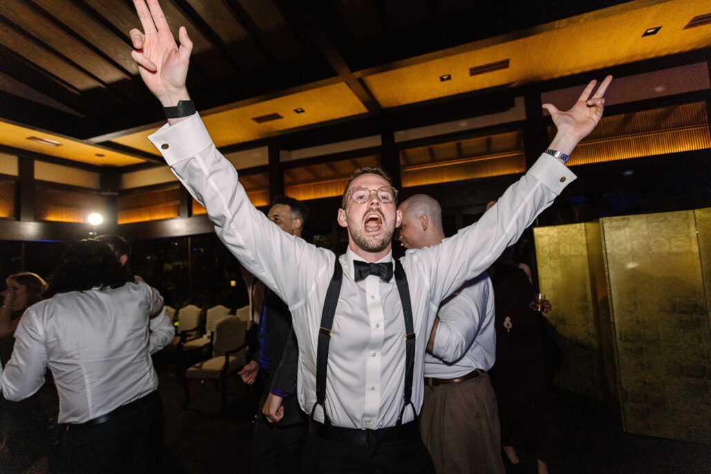 A groomsman in braces throws his arms wide, cheering.