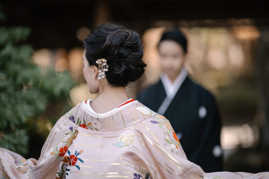 Behind the bride's hair ornament and uchikake collar, the groom in black montsuki stands soft in the background.
