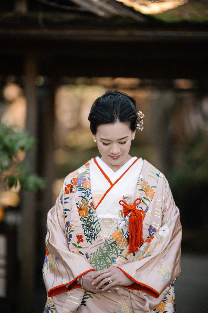 The bride in her uchikake stands beneath a wooden gate, head bowed, the red lining showing at her collar.
