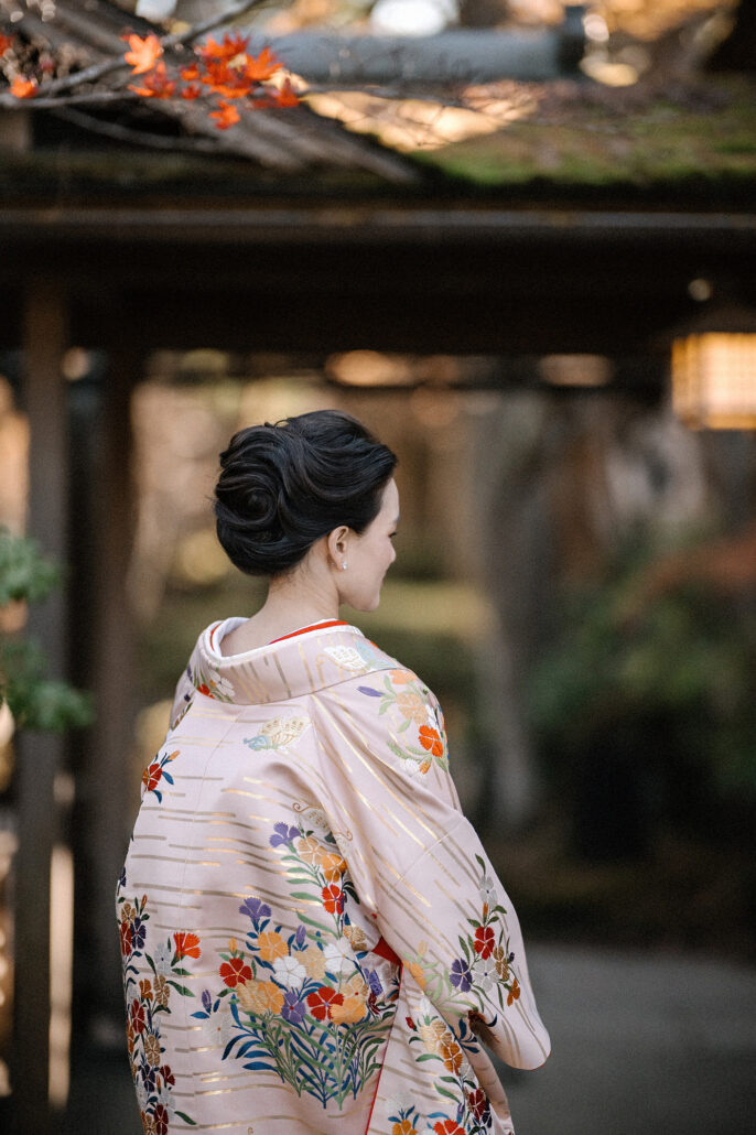 The back of the bride's uchikake, flowers and gold bands across the silk, as she looks aside under autumn leaves.