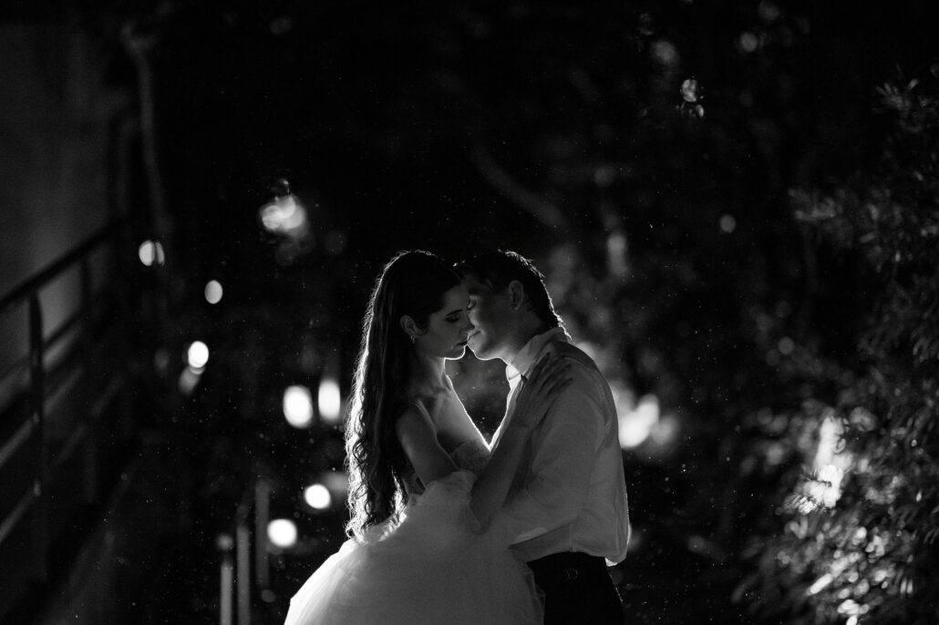 Black and white photograph of a bride and groom embracing at night, lanterns glowing in the garden behind them