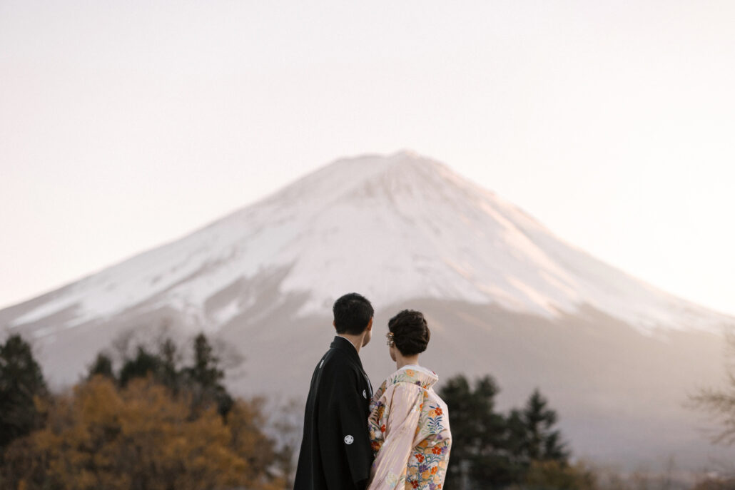 Seen from behind, the bride and groom in formal kimono look toward Mt Fuji, its summit clear in the last light.