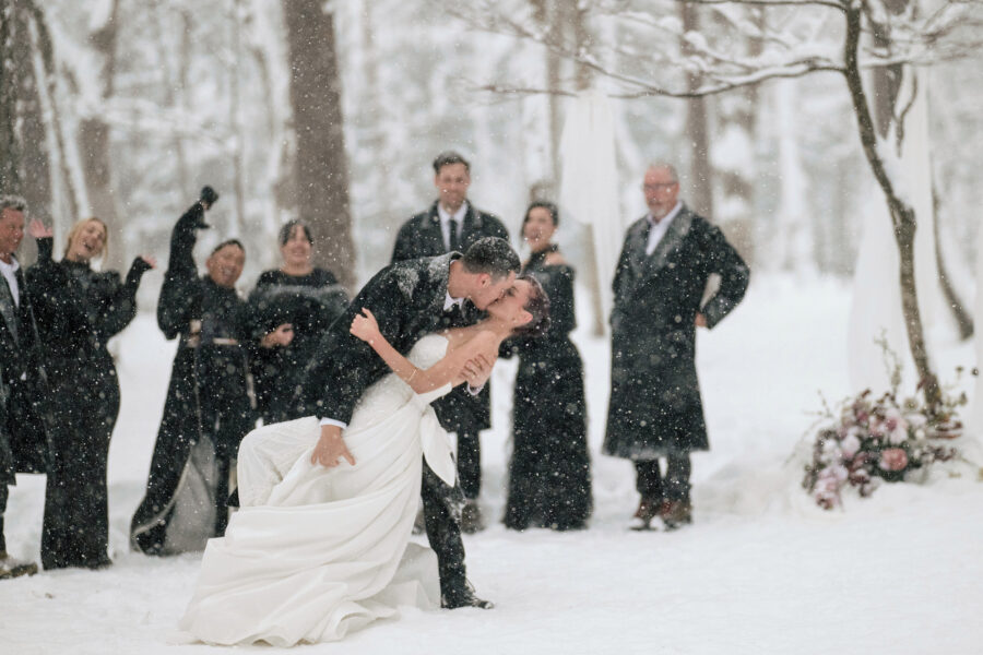 The groom dips the bride for a kiss in heavy falling snow as the wedding party cheers in the forest at their Hakuba snow wedding.
