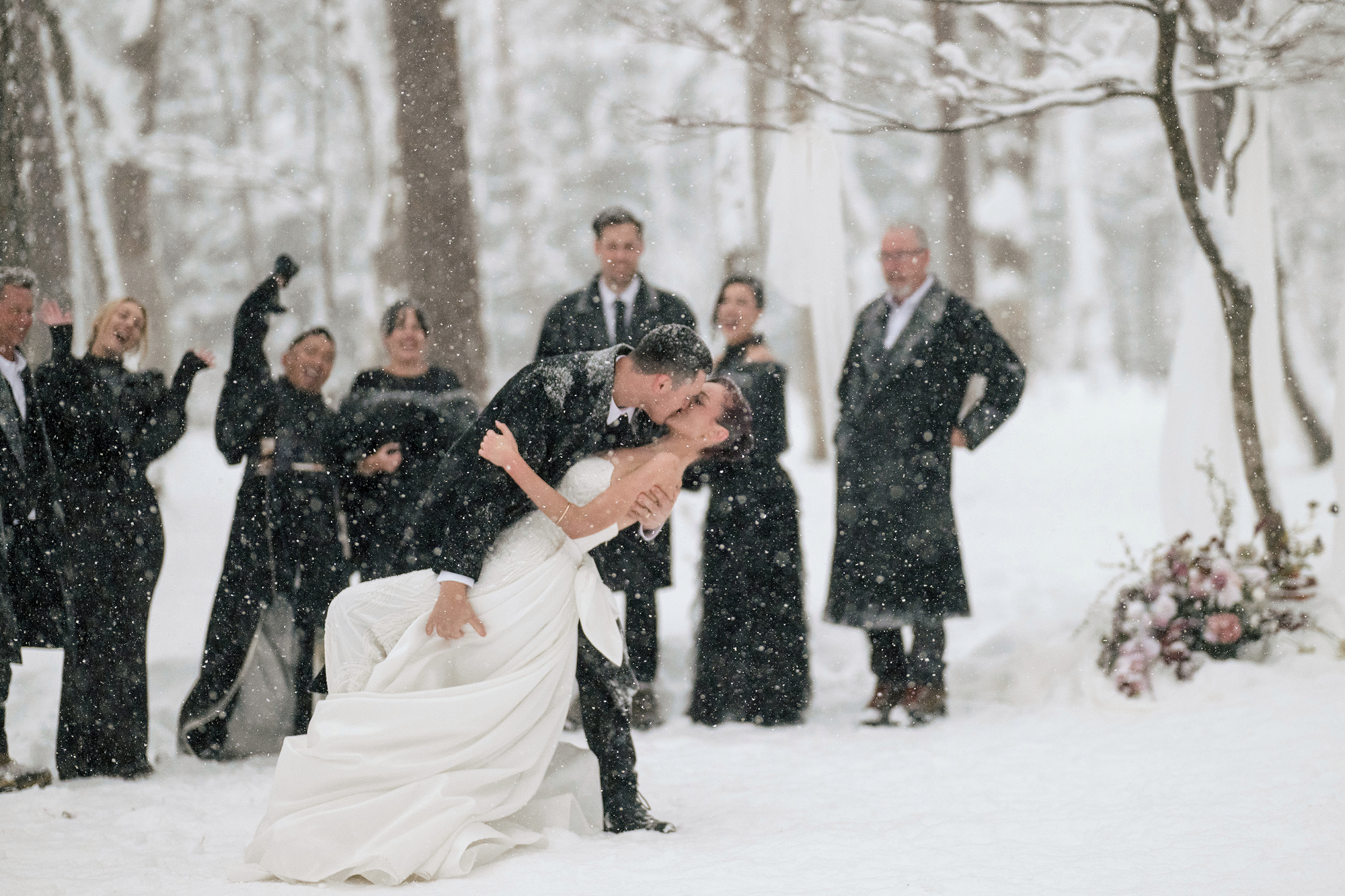 The groom dips the bride for a kiss in heavy falling snow as the wedding party cheers in the forest at their Hakuba snow wedding.