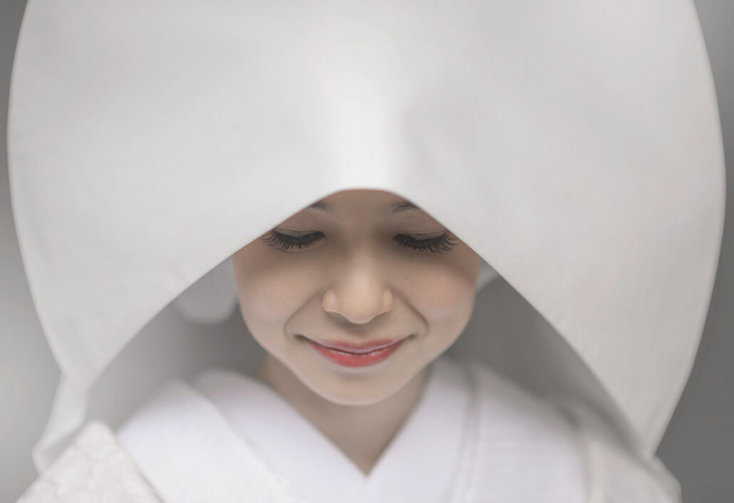 Under a white wataboshi hood, a Japanese bride looks down with a quiet smile, her red lips the only colour in the frame.