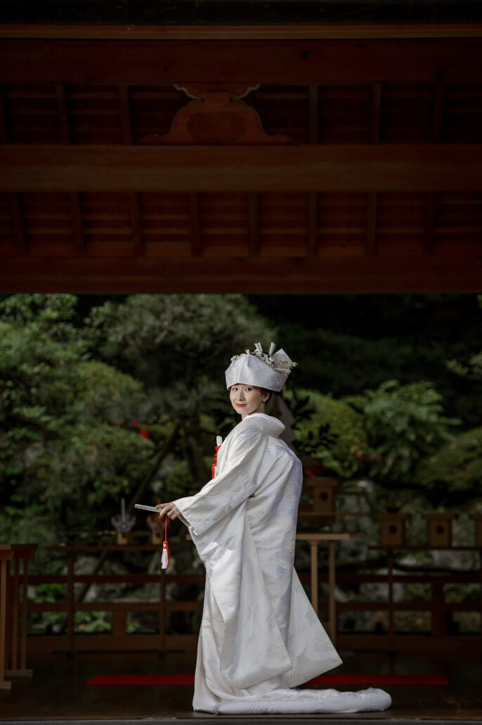 A bride in a white tsunokakushi and crane-patterned shiromuku glances back from the vermilion noh stage where her Shinto wedding took place.