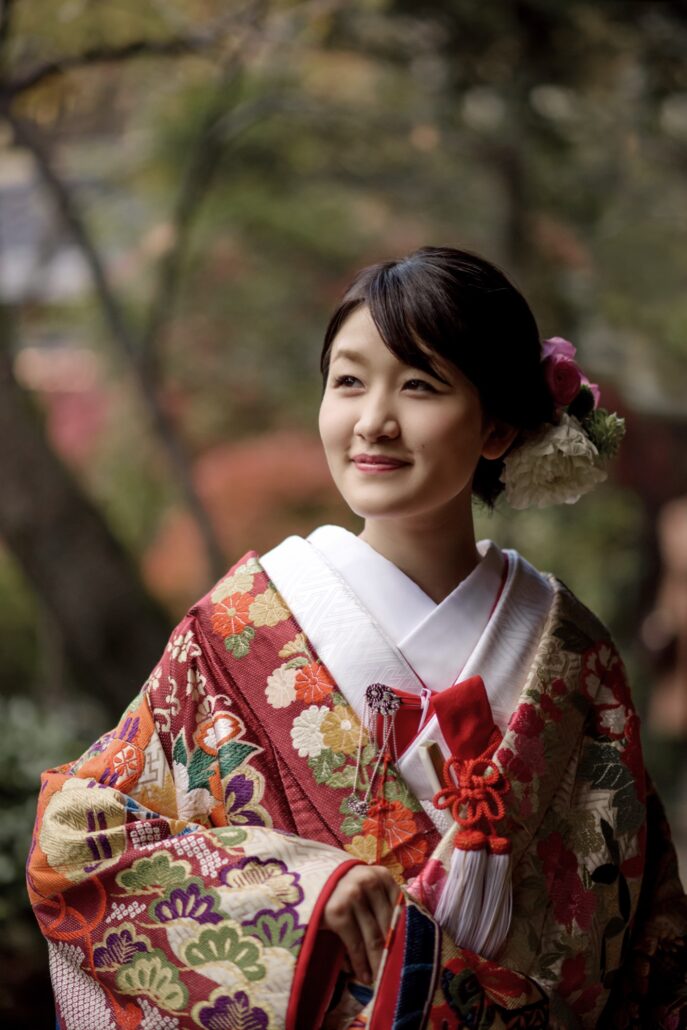 A bride in a red and gold iro-uchikake patterned with chrysanthemums and fans, a tasselled red cord at her chest, in an autumn garden.