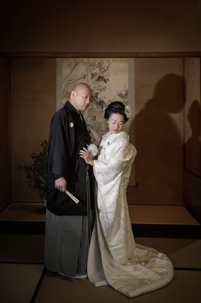 A bride with traditional nihongami hair, her white uchikake trailing over tatami, stands close to her groom beside a hanging scroll.