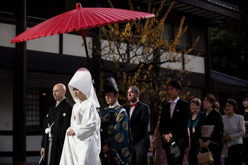 Under a red umbrella, a bride in a white wataboshi and her groom in montsuki lead both families in a Shinto wedding procession.