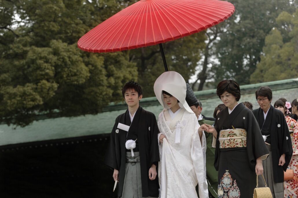 A bride in a white wataboshi walks beside her groom under a red umbrella, an older woman in a black kurotomesode kimono at her side.