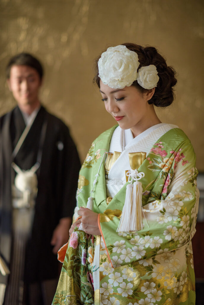 A bride in a pale green and gold iro-uchikake with cherry blossoms, white peonies in her hair, as her groom in montsuki waits behind.