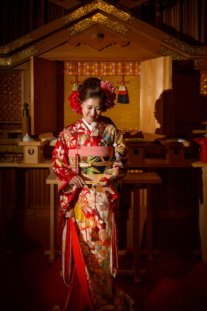 A bride in a red hikifurisode with a tied obi and a long trailing hem, red flowers in her hair, stands before a Shinto altar.
