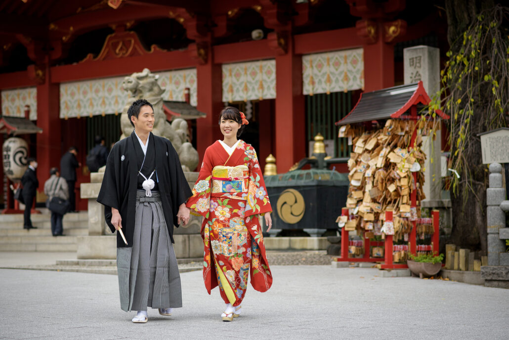 Hand in hand, a bride in a red furisode with a tied obi and her groom in montsuki and hakama walk past ema plaques at a vermilion shrine.