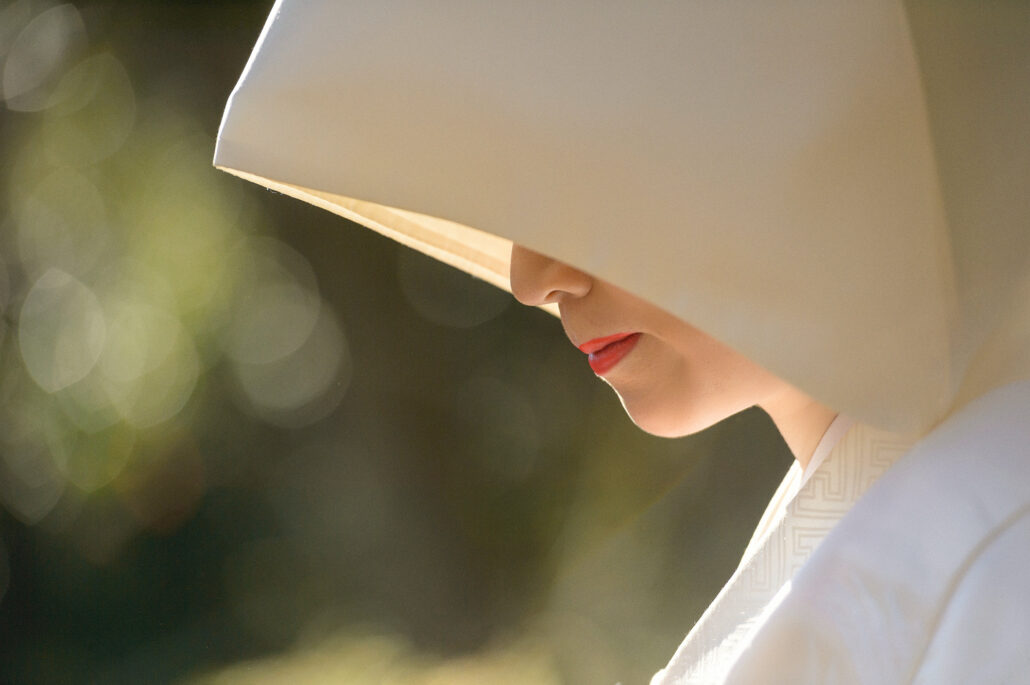 In profile, a bride's face is half hidden by the brim of her white wataboshi, only her red lips catching the winter garden light.