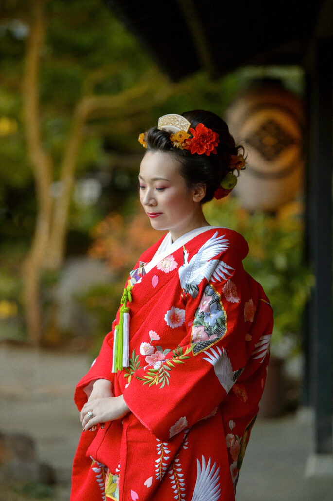 A bride in a red iro-uchikake embroidered with white cranes and cherry blossoms, a gold kanzashi comb and red flowers in her hair.