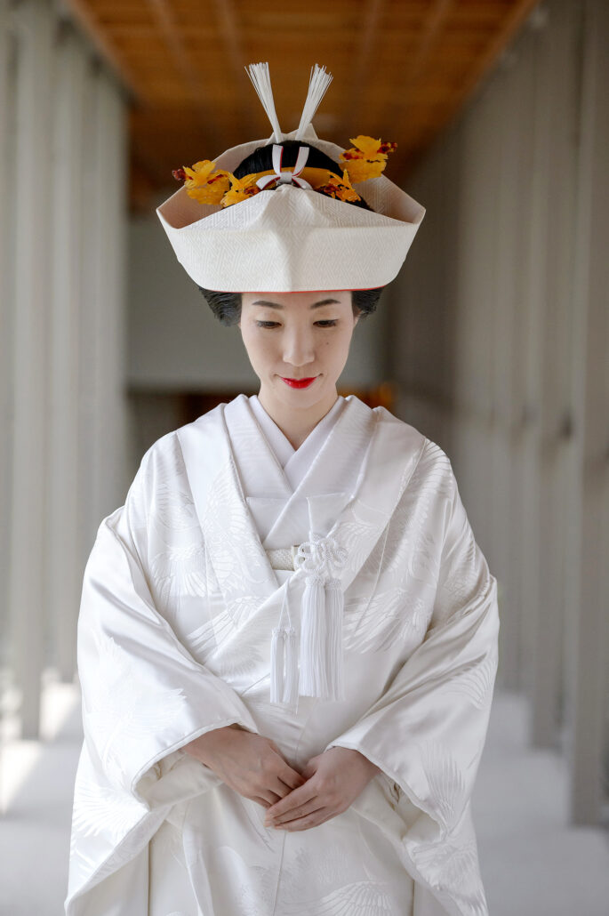 A bride in a white tsunokakushi headdress with gold ornaments and a crane-patterned shiromuku kimono stands with hands folded.