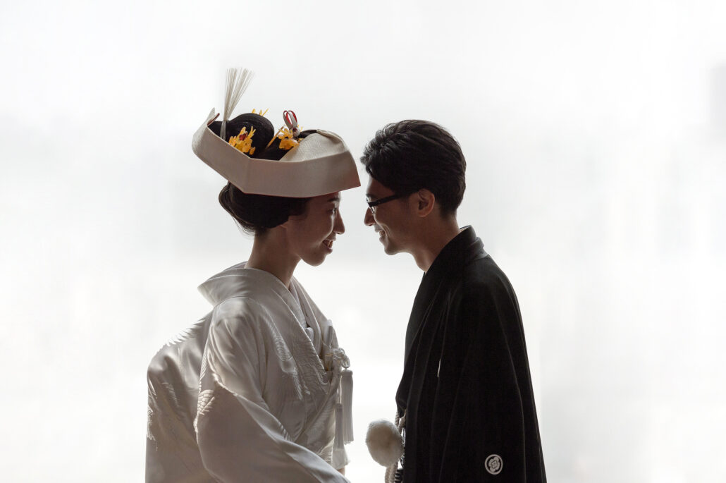 Foreheads almost touching, a bride in a tsunokakushi and white crane shiromuku smiles at her groom in black montsuki against bright window light.