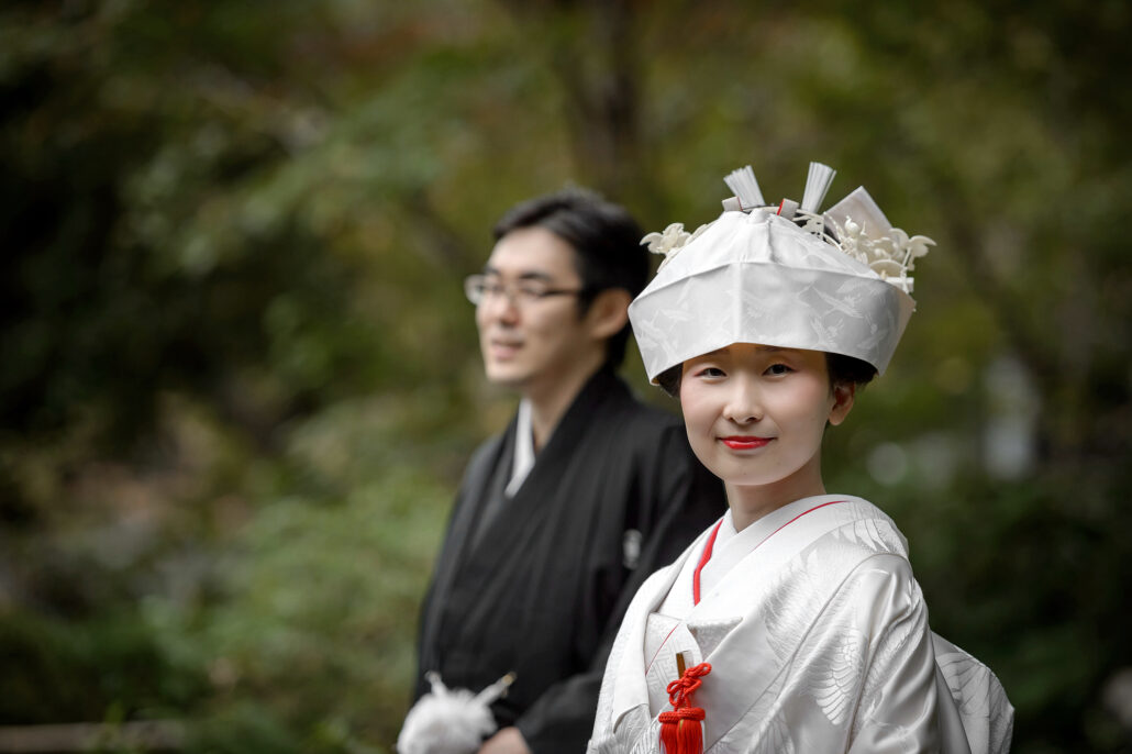A bride in a white tsunokakushi and crane shiromuku, a red cord at her chest, smiles at the camera as her groom stands beside her.