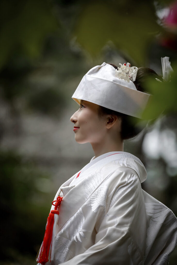 In profile against dark garden green, a bride in a white tsunokakushi headdress and shiromuku kimono, a red tassel at her chest.