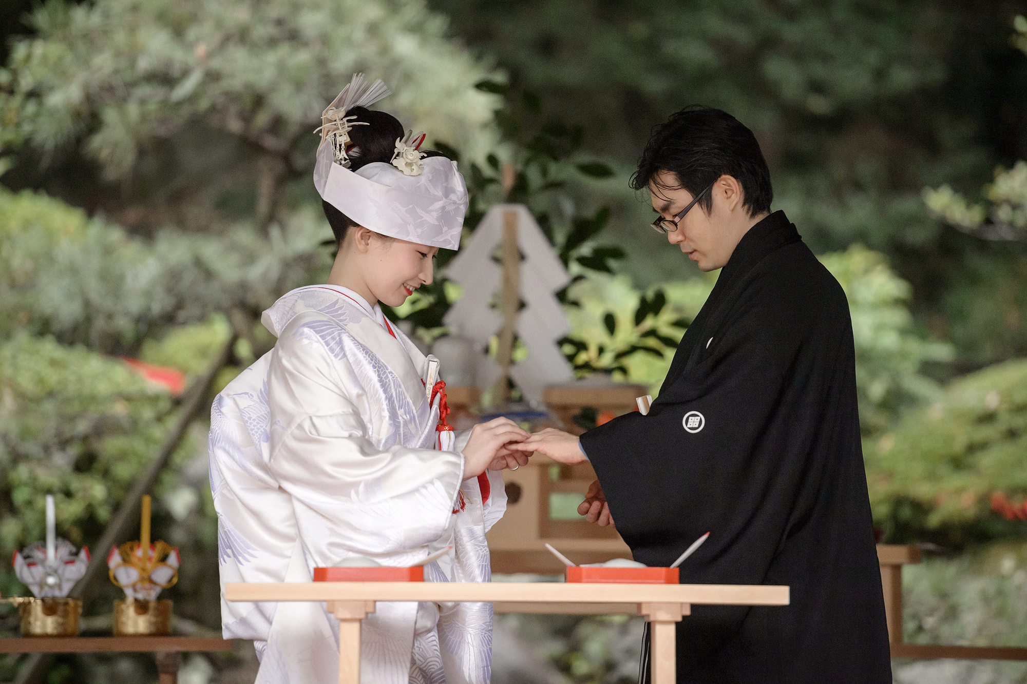 The groom in black montsuki places the ring on the bride's finger before the altar at a Shinto shrine wedding in a Japanese garden.