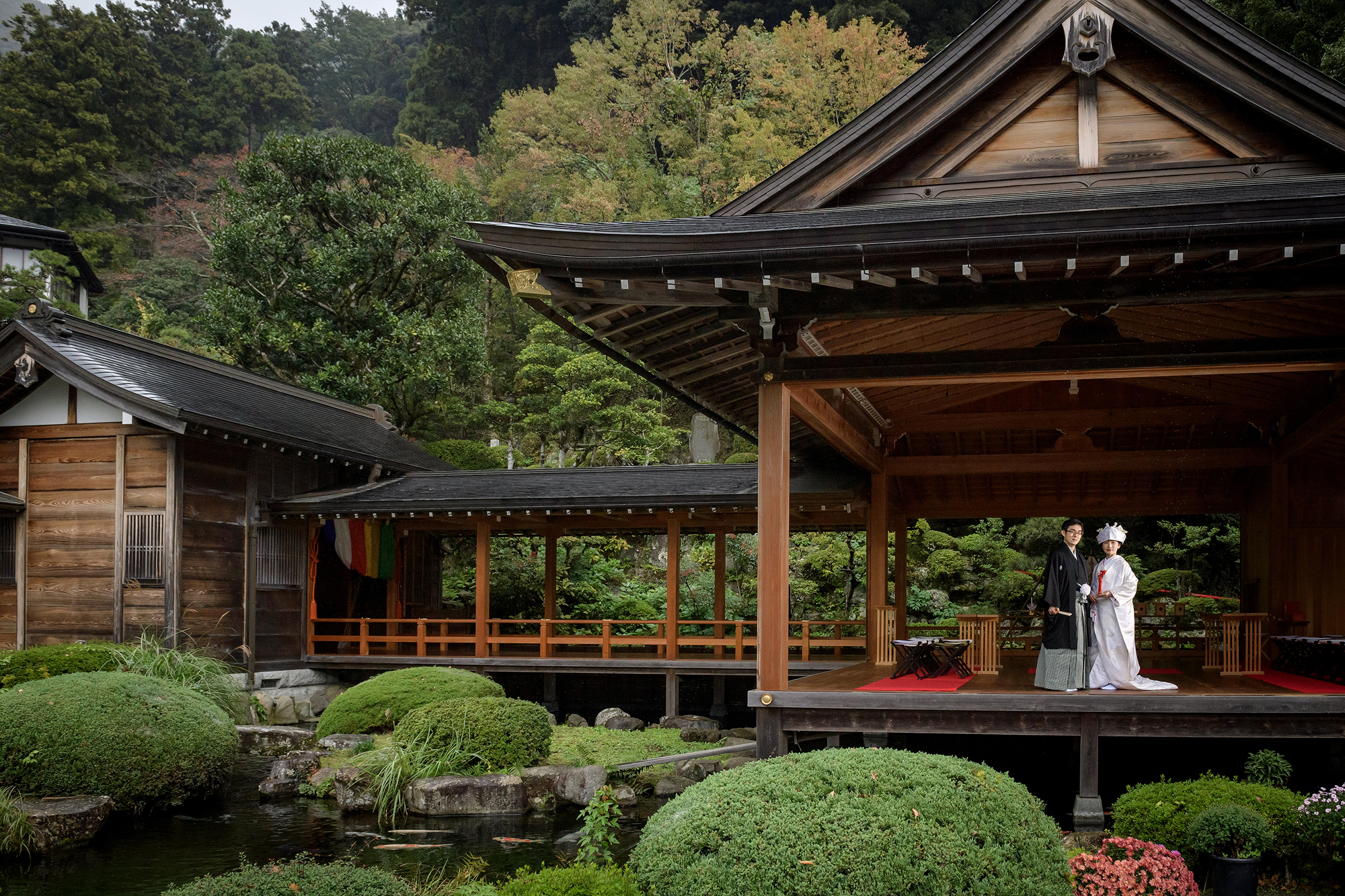 The bride and groom in wedding kimono stand small on the old stage in the garden at Jinya, clipped azaleas and a curved roof around them, after their Shinto wedding in Japan.