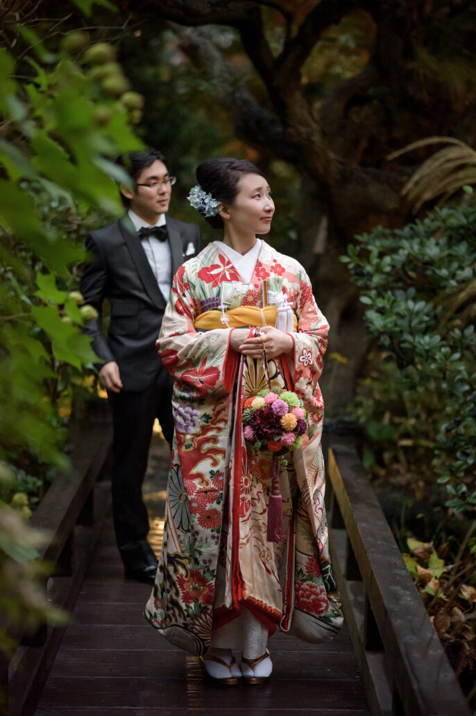 After changing from white, the bride in a cream and red hikifurisode with a tied obi walks a garden path, her groom in black tie behind.