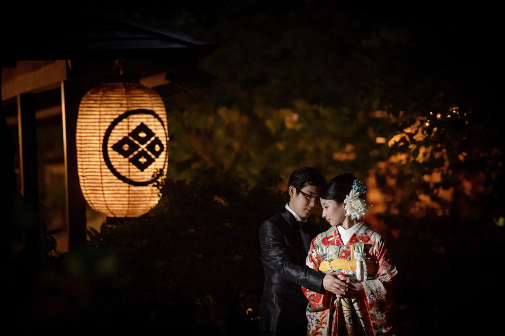 At dusk, a bride in a red and cream kimono leans into her groom beside a glowing paper lantern carrying the venue's crest.