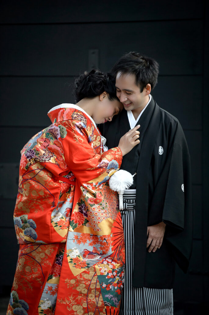 A bride in a red and gold iro-uchikake rests her head on her groom's shoulder as he smiles in montsuki and striped hakama.