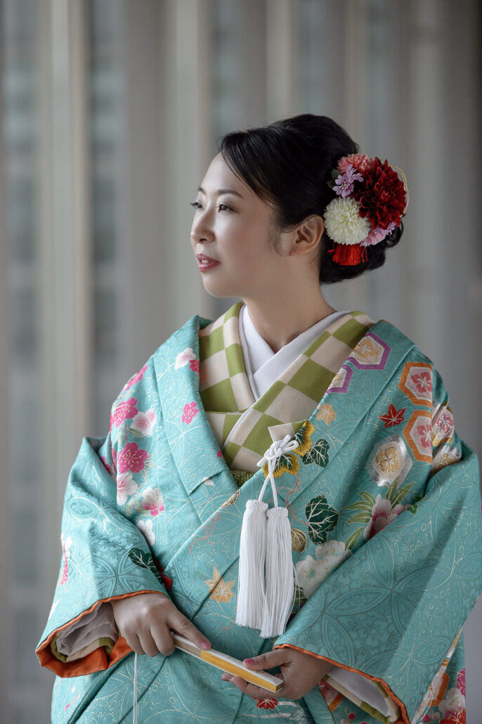 A bride in a turquoise iro-uchikake with cherry blossoms and a green checked collar holds a folded fan, red and white flowers in her hair.