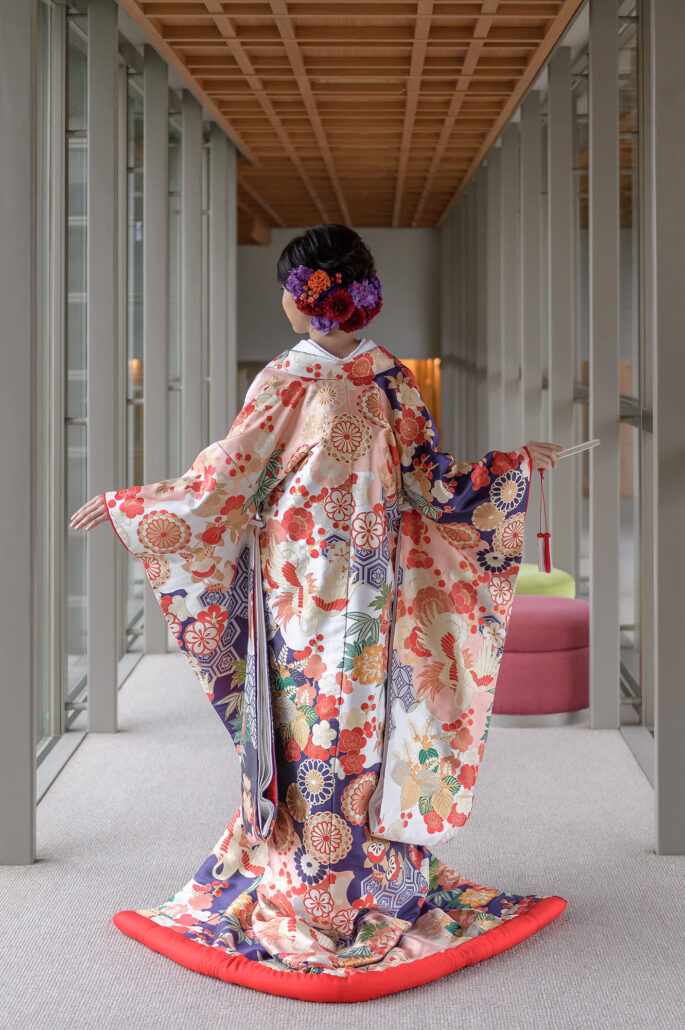 Seen from behind with arms outstretched, a bride shows the full shape of her iro-uchikake, cranes and chrysanthemums above a padded red hem.