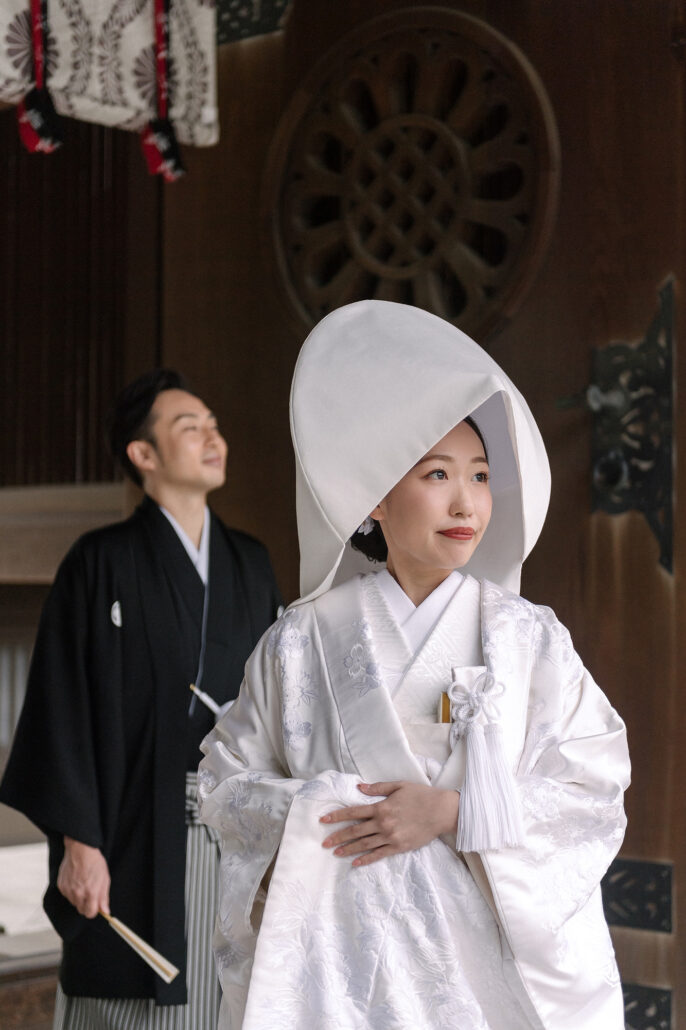 At the Meiji Jingu shrine hall in Tokyo, a bride in a white wataboshi and shiromuku stands with her groom in montsuki behind her.
