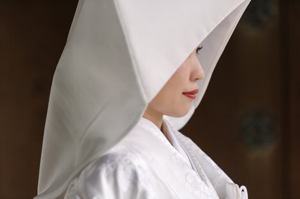 A close profile of a bride under the folded white silk of her wataboshi, red lips just visible below the brim, Tokyo.
