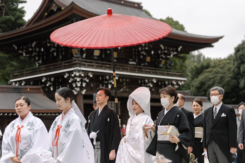 A Shinto wedding procession crosses the Meiji Jingu courtyard under a red umbrella, shrine maidens leading the couple and their families.