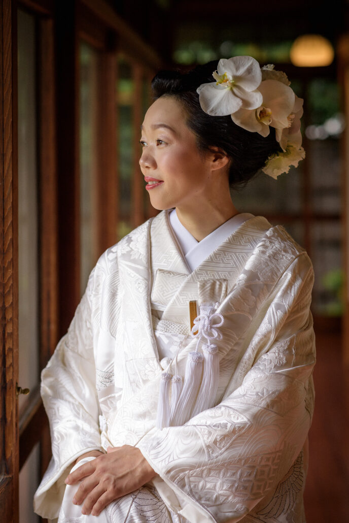 A bride in a white uchikake, white orchids in her traditional nihongami hair, sits by a wooden window looking out at the garden.