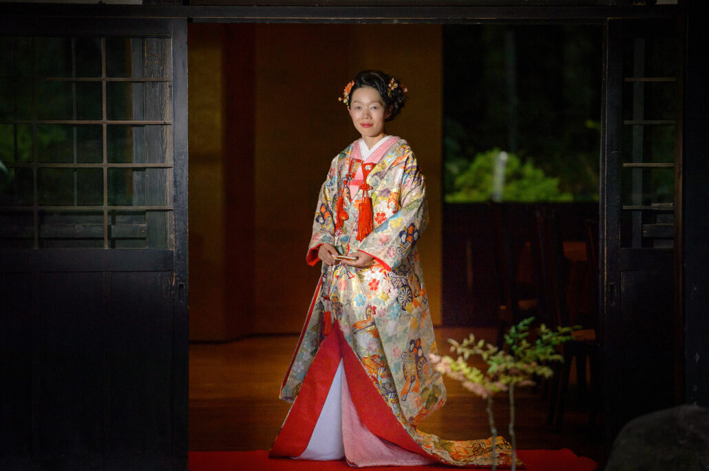 Framed by a dark wooden doorway, a bride stands in a full-length iro-uchikake, its padded red lining and hem spread on the floor.