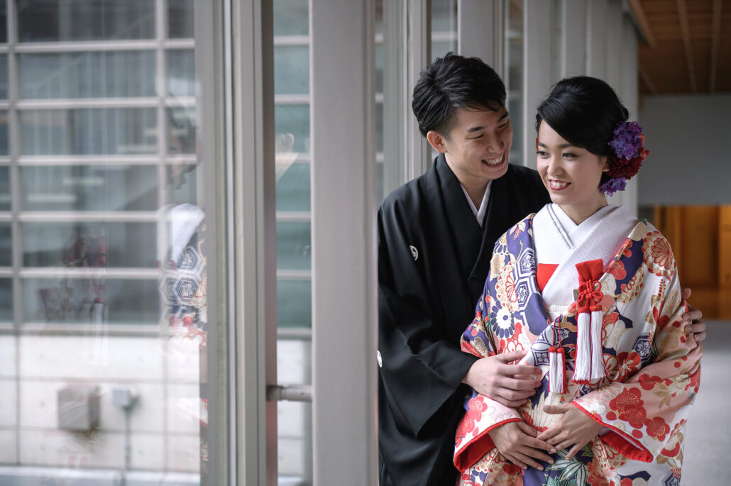 Laughing together by tall windows, a bride in a red and navy iro-uchikake leans back into her groom in black montsuki.