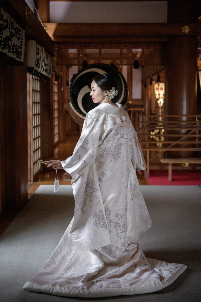 Inside a shrine hall, a bride turns in a white uchikake embroidered with cranes and blossoms, the great shrine drum behind her.
