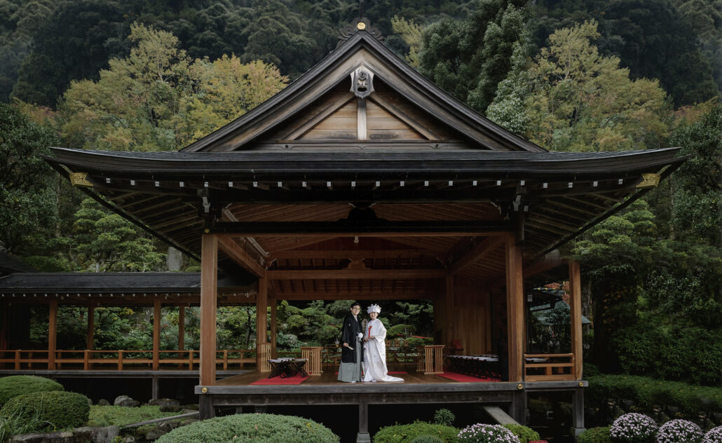 A wide view of a Shinto wedding on a garden noh stage, the bride in white and her groom small beneath its great wooden roof, clipped pines and azaleas all around.