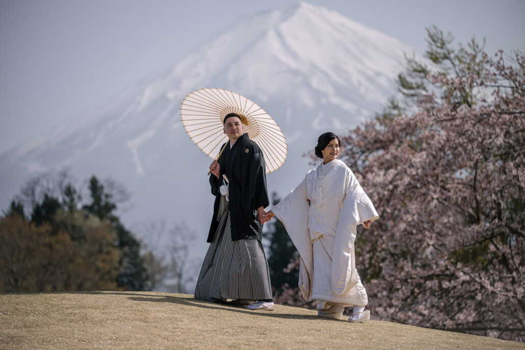 Beneath Mt Fuji and spring cherry blossom, a bride in a white shiromuku walks beside her groom in montsuki and hakama holding a paper umbrella.