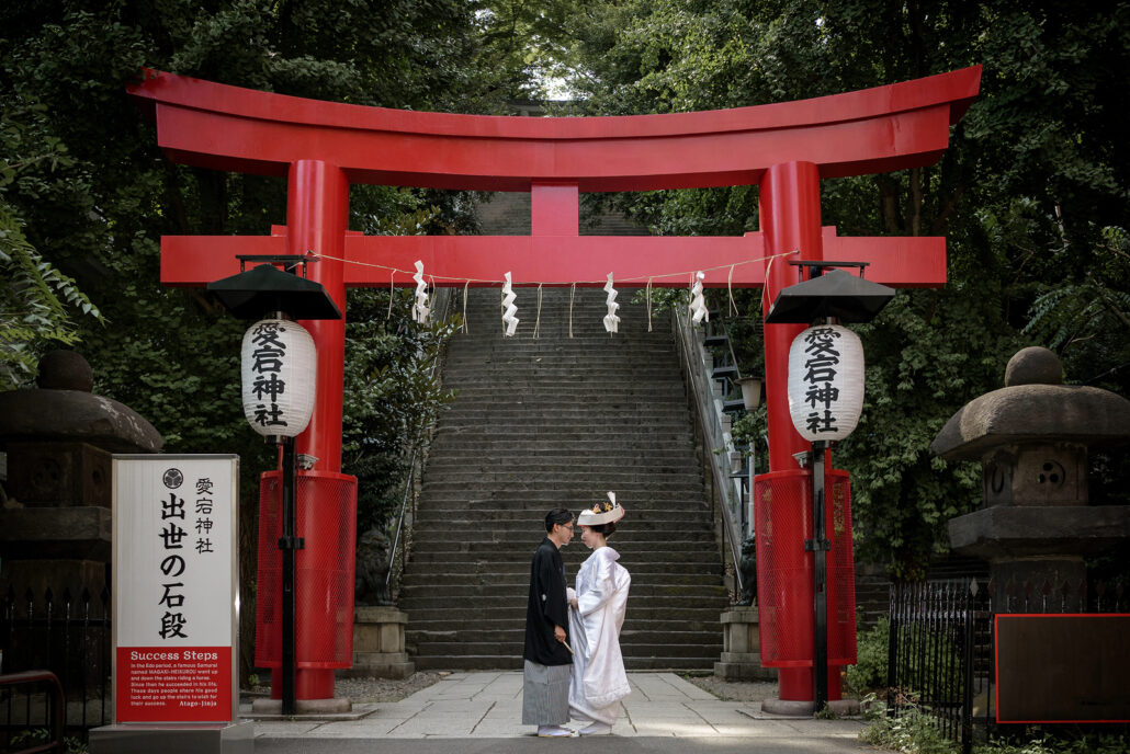 Beneath the vermilion torii of Atago Shrine in Tokyo, a bride in a tsunokakushi and white shiromuku faces her groom at the foot of the steep stone steps.