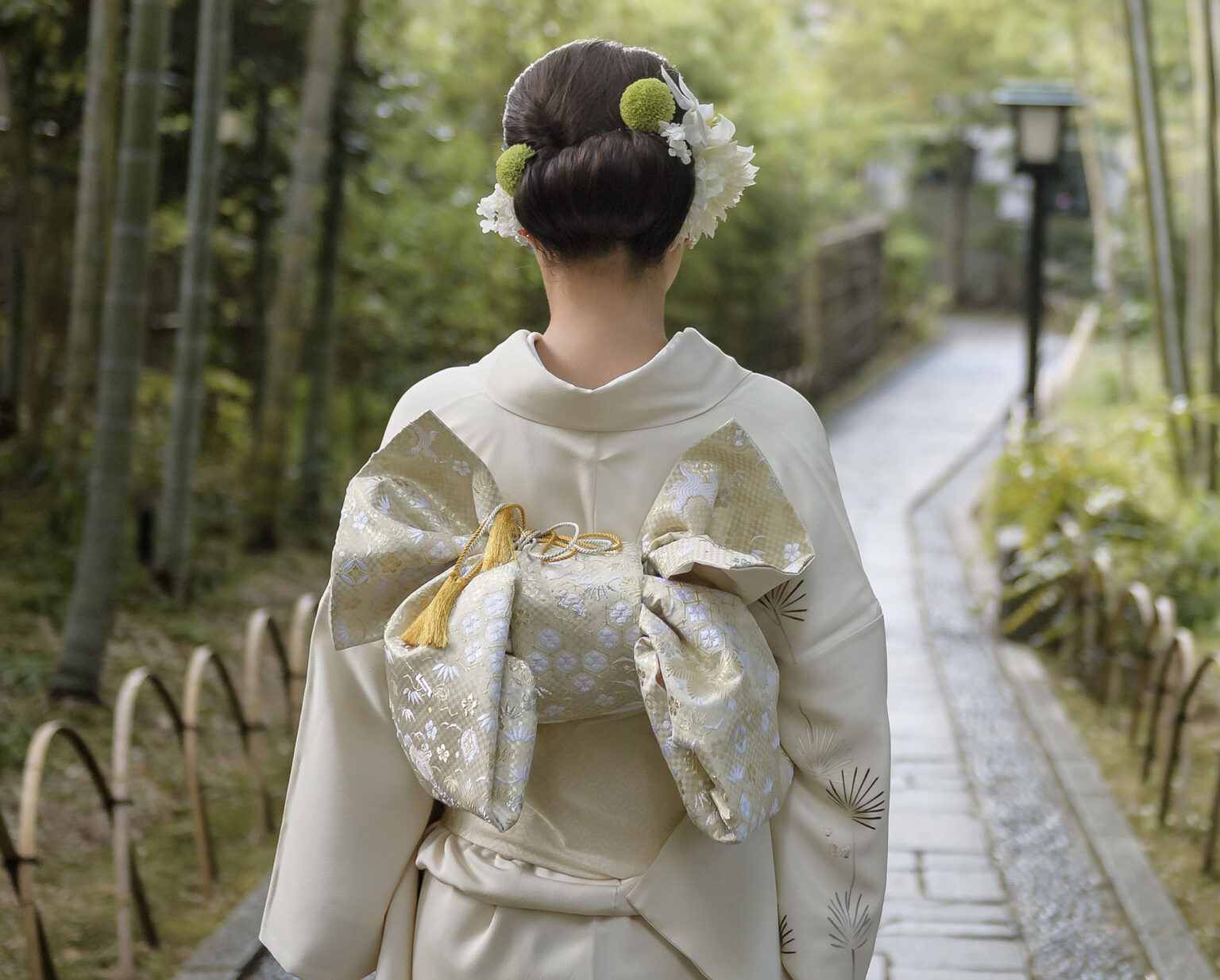 Seen from behind on a stone path through bamboo in Shuzenji, a bride in an ivory kimono walks toward a lantern, her gold obi tied in a full bow.