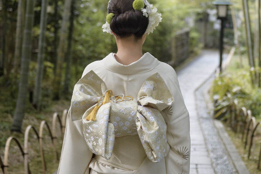 Seen from behind on a stone path through bamboo in Shuzenji, a bride in an ivory kimono walks toward a lantern, her gold obi tied in a full bow.