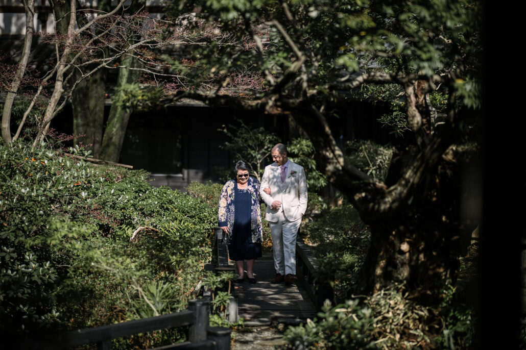 The groom walks an older woman in a floral dress along a garden path toward the ceremony, maples arching overhead