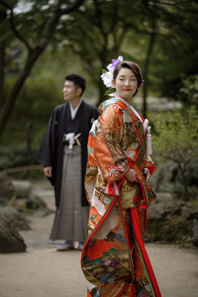 In a green garden, a bride in an orange and gold iro-uchikake embroidered with white cranes looks back over her shoulder, her groom in hakama behind.