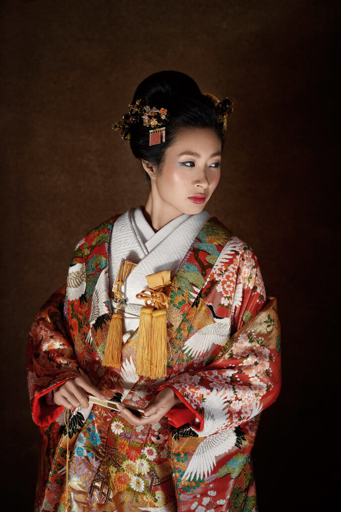 A studio portrait of a bride in a red iro-uchikake with white cranes and cherry blossom, gold kanzashi in her traditional hair, a fan in her hands.