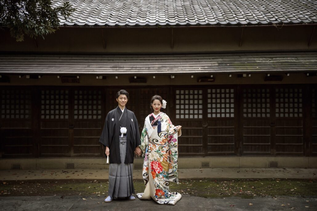 Hand in hand before an old wooden hall, a groom in montsuki and hakama and a bride in a white iro-uchikake painted with peonies and swirling water.