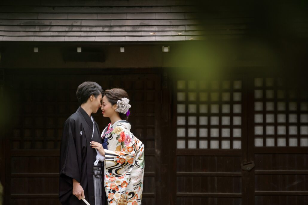 Foreheads touching, a bride in a white iro-uchikake painted with flowers and swirling water leans into her groom in black montsuki.