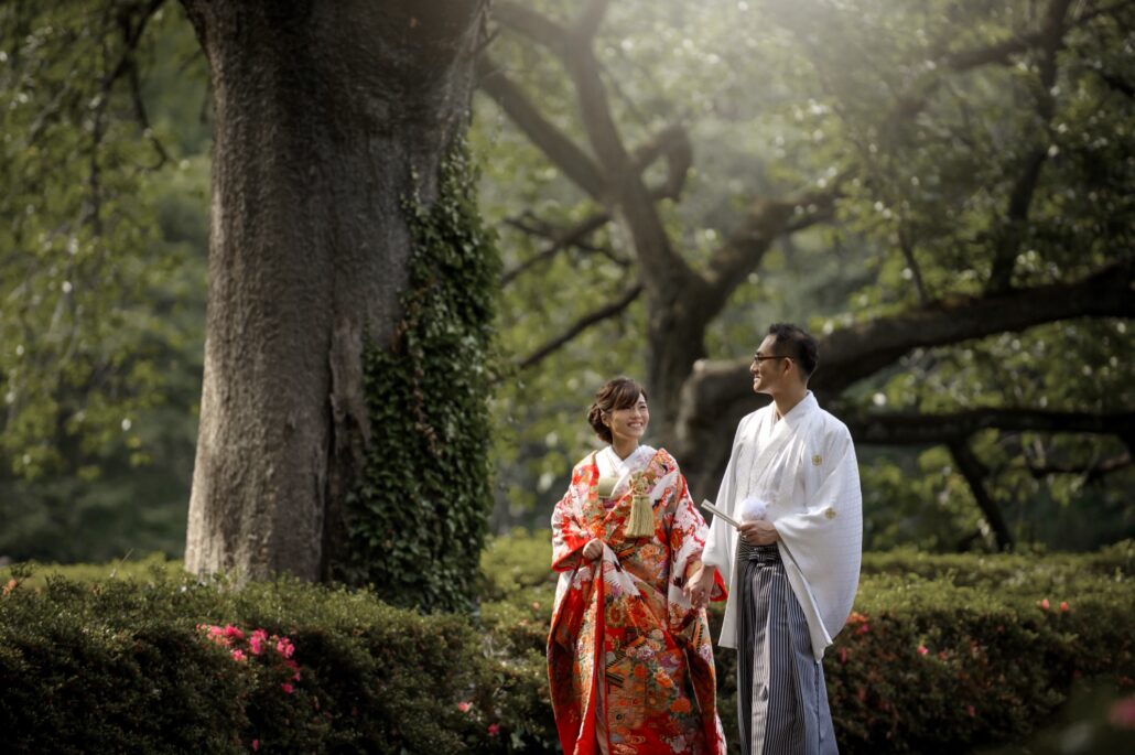 Under a great ivy-covered tree, a bride in a red iro-uchikake with white cranes smiles at her groom in a white montsuki and striped hakama.