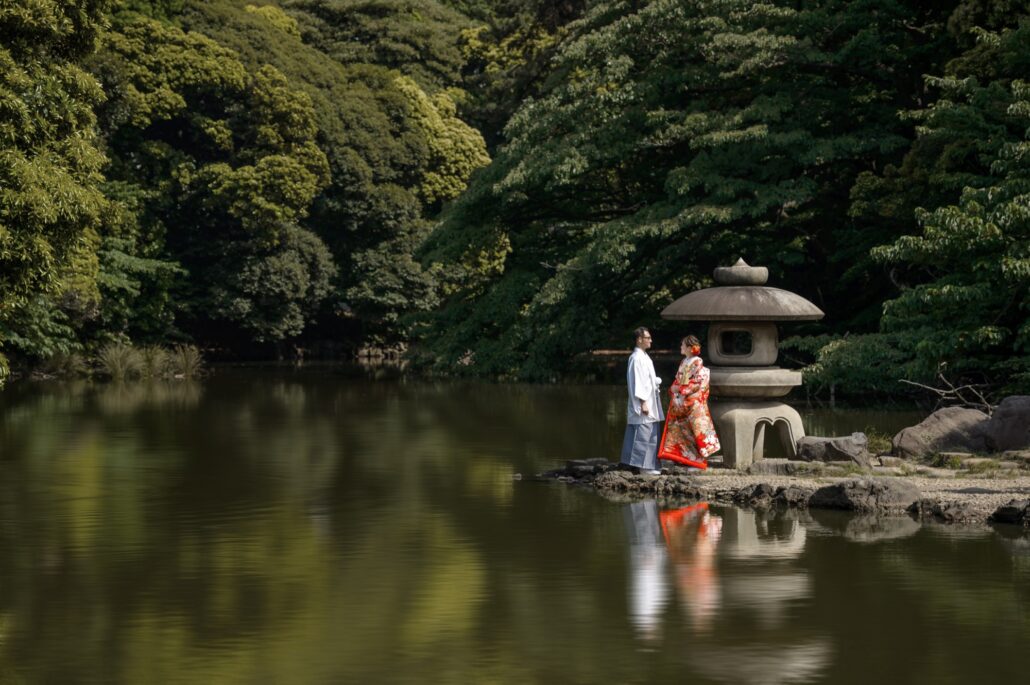 Beside a stone lantern on a Tokyo garden pond, a bride in a red iro-uchikake and her groom in a white montsuki are mirrored in the still water.
