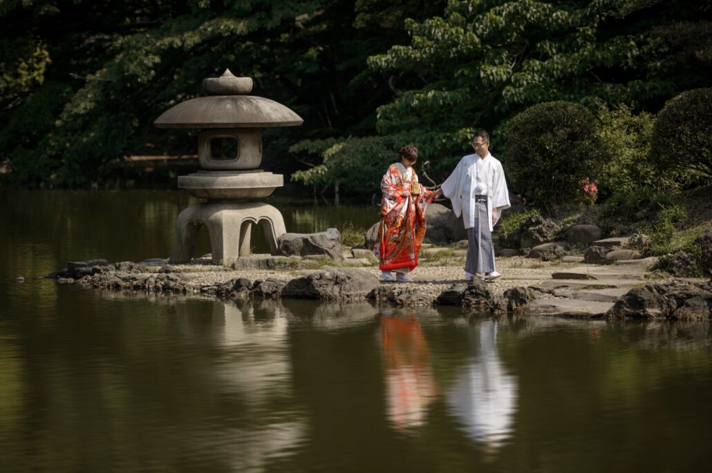 Holding hands, a bride in a red iro-uchikake and her groom in a white montsuki walk along a pond edge past a stone lantern in Tokyo.